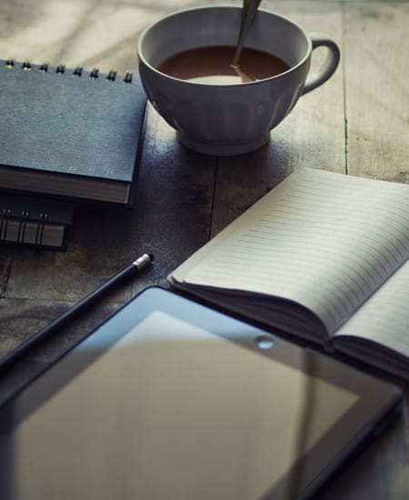 Moody photo of a desk with a tablet, an opened blank notebook, a pencil, closed notebooks, and a half-filled cup of coffee on it.